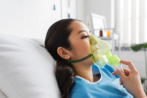 side view of sick asian woman in oxygen mask lying on bed in hospital ward