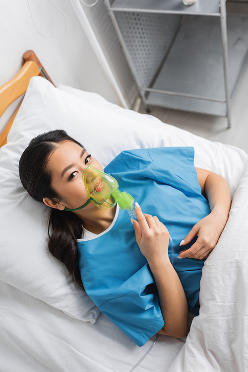 top view of positive asian woman breathing in oxygen mask and looking at camera on bed in clinic