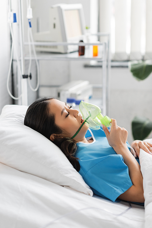 side view of sick asian woman holding oxygen mask while lying on bed in hospital ward