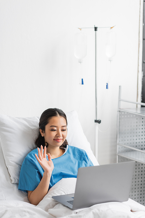 cheerful asian woman waving hand during video chat on laptop in hospital ward