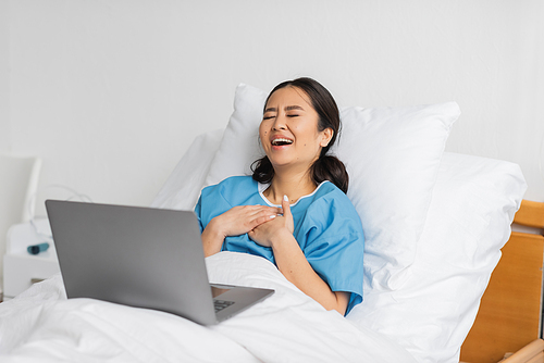 excited asian woman touching chest and laughing with closed eyes while watching comedy film on laptop in clinic