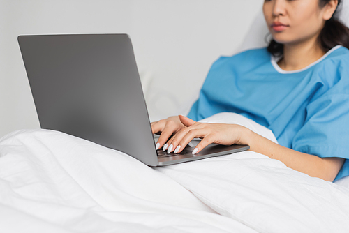 partial view of asian woman typing on laptop on bed in hospital ward
