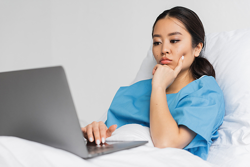 thoughtful asian woman lying on hospital bed and looking at laptop