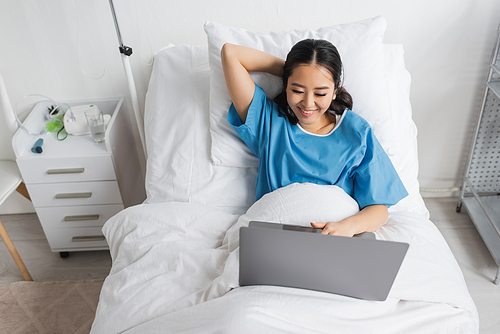 high angle view of cheerful asian woman using laptop on bed in clinic