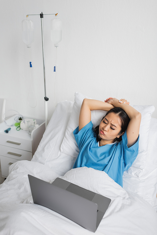 displeased asian woman looking at laptop while watching movie in hospital ward