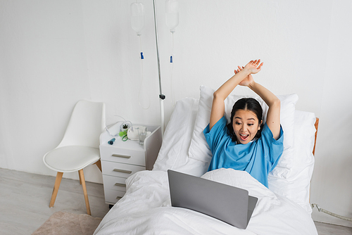 excited asian woman with raised hands watching movie on laptop in hospital ward