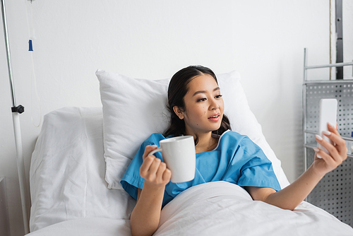 surprised asian woman with cup of tea looking at mobile phone in hospital ward