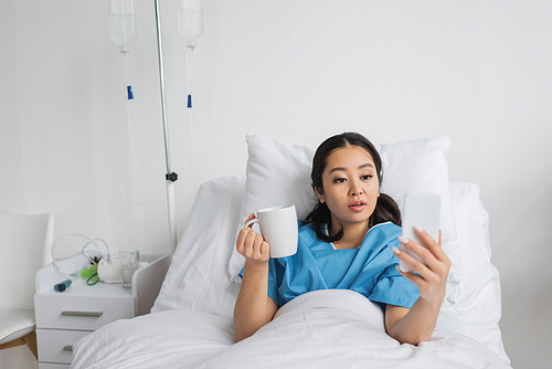 amazed asian woman holding cup of tea and looking at mobile phone on bed in clinic