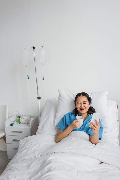 excited asian woman with smartphone and cup of tea laughing with closed eyes on hospital bed