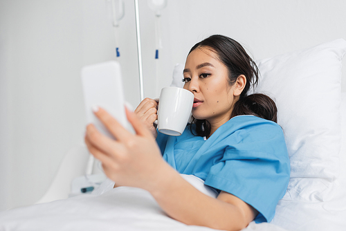 young asian woman drinking tea and looking at blurred cellphone in hospital ward