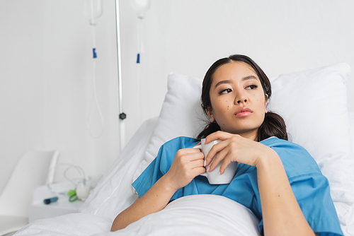 thoughtful asian woman holding tea cup and looking away on hospital bed