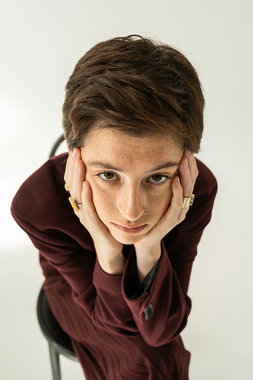 high angle view of thoughtful freckled woman in blazer and finger rings holding hands near face and looking at camera on grey