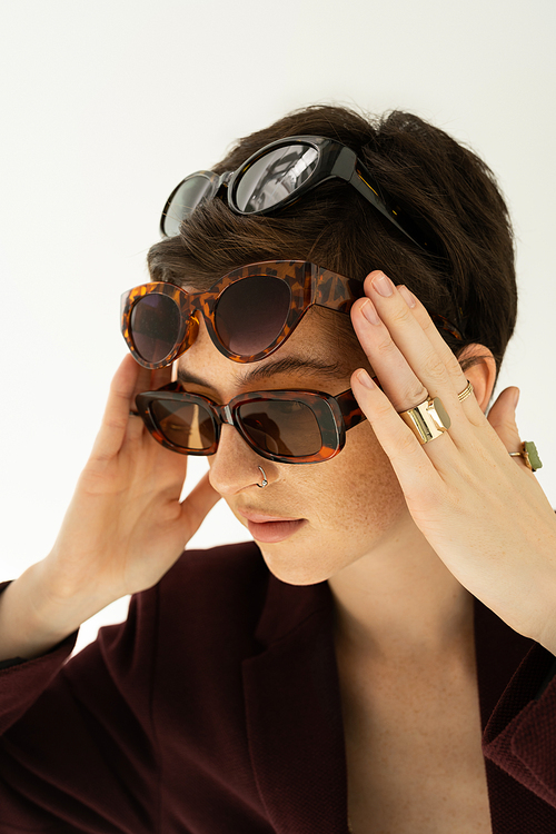 young woman in finger rings trying on several stylish sunglasses isolated on grey