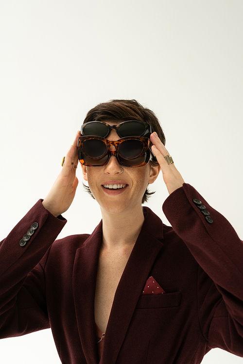 cheerful woman in burgundy brown blazer fitting on several stylish sunglasses isolated on grey