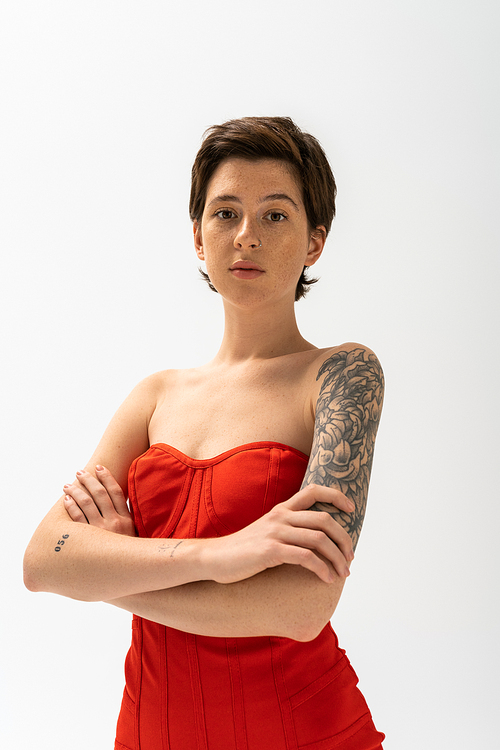 young tattooed woman in red strapless dress crossing arms and looking at camera on grey background