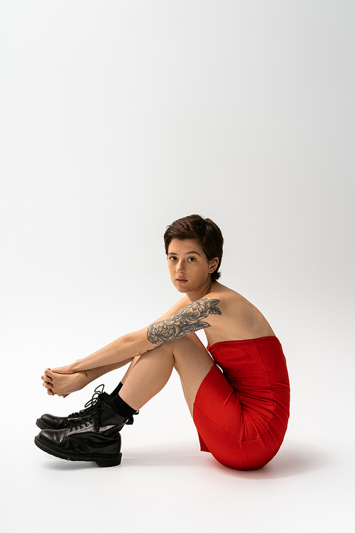 full length of brunette tattooed woman sitting in red corset dress and rough boots on grey background