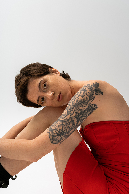 dreamy tattooed woman in red corset dress sitting and looking at camera on grey background