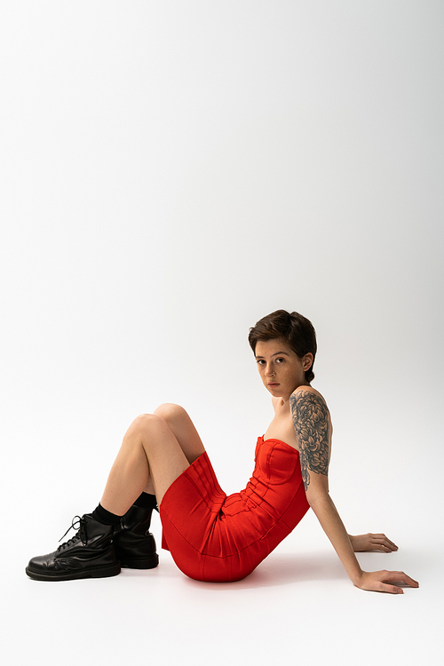 young tattooed woman in black rough boots and red corset dress sitting and looking at camera on grey background