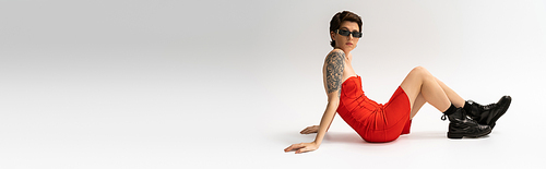 full length of slim tattooed woman in black boots and red corset dress sitting on grey background, banner