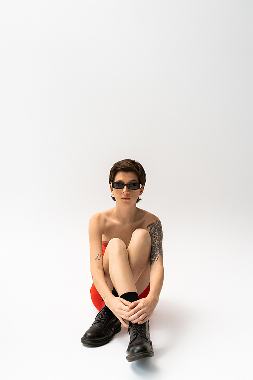 full length of young tattooed woman in black rough boots and trendy sunglasses sitting on grey background