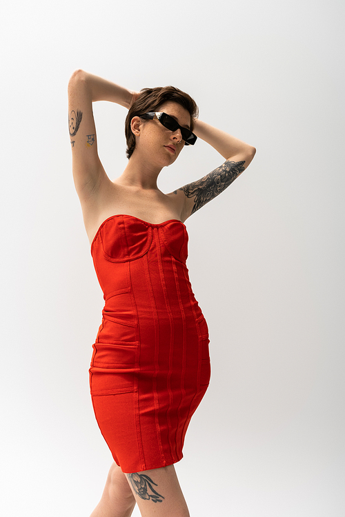 low angle view of tattooed woman in sunglasses and red corset dress posing with hands behind head on grey