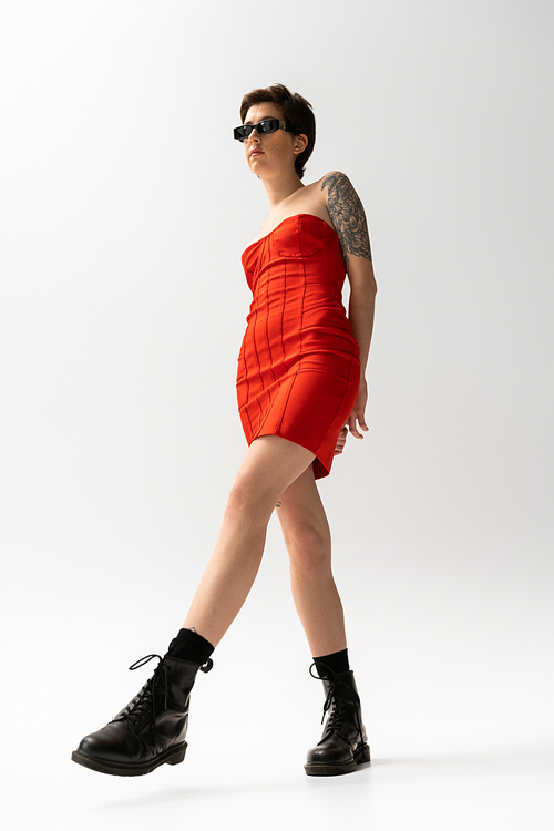 full length of slender tattooed woman posing in red corset dress and black boots on grey background