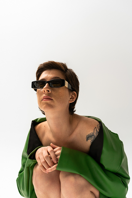 tattooed woman in green leather jacket and fashionable sunglasses sitting on grey background