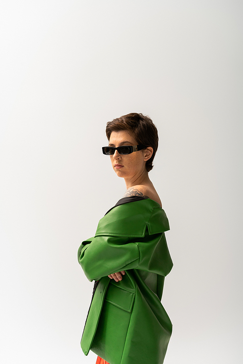 brunette tattooed woman in green leather jacket and stylish sunglasses looking at camera isolated on grey