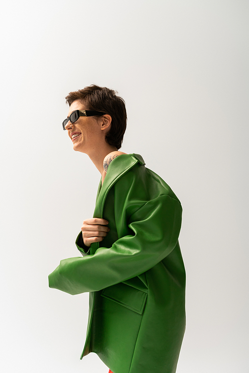 side view of overjoyed brunette woman posing in trendy sunglasses and green leather jacket isolated on grey