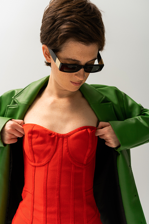 stylish woman in sunglasses and green leather jacket adjusting red strapless dress isolated on grey