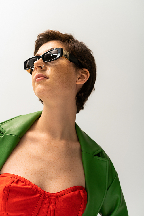 low angle view of young brunette woman in sunglasses and green leather jacket looking away isolated on grey