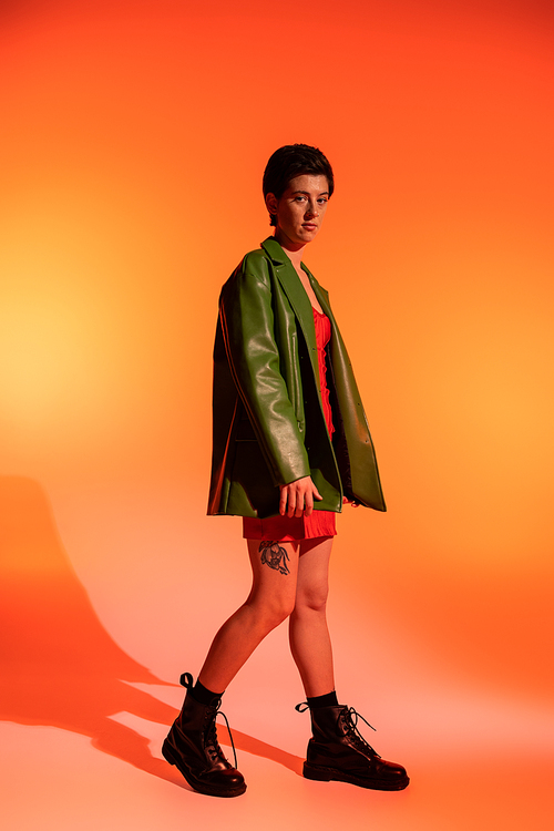 full length of tattooed woman in green leather jacket and black boots looking at camera on orange background