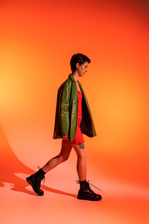 side view of slender woman in black boots and green leather jacket walking on orange background
