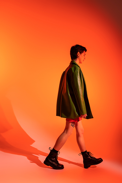side view of tattooed woman in green jacket and black boots walking on orange background