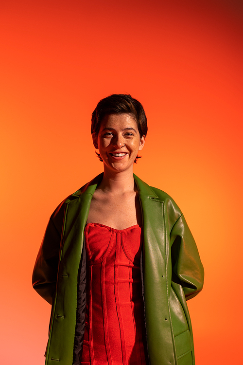 carefree brunette woman in green leather jacket and red corset dress looking at camera on orange background