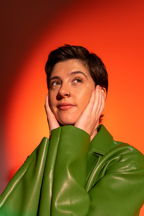 dreamy and positive woman in green leather jacket touching face and looking away on orange shaded background