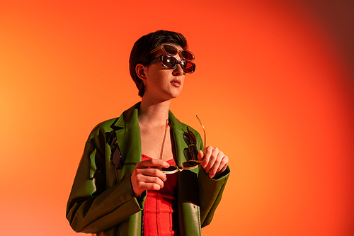 trendy woman in green leather jacket posing in several sunglasses and looking away on orange background
