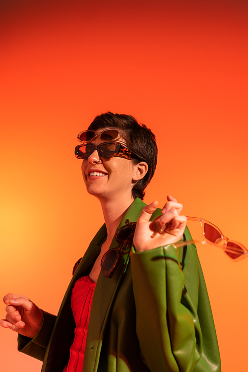 young and carefree woman posing in green stylish jacket and different trendy sunglasses on orange background