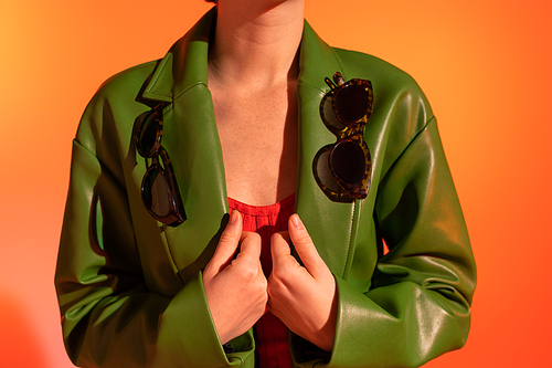 partial view of woman with stylish sunglasses on green leather jacket on orange background