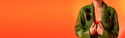 cropped view of woman posing with trendy sunglasses on green leather jacket on orange background, banner