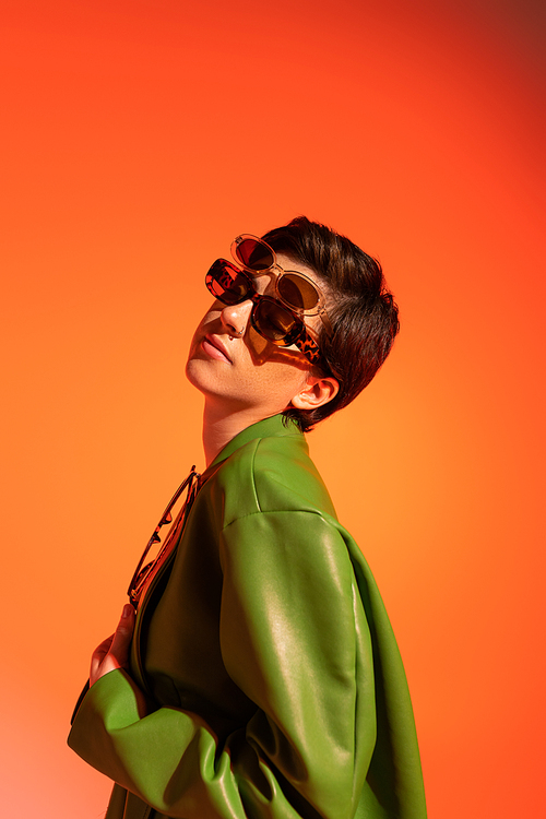 stylish brunette woman posing in several sunglasses and green leather jacket on orange background
