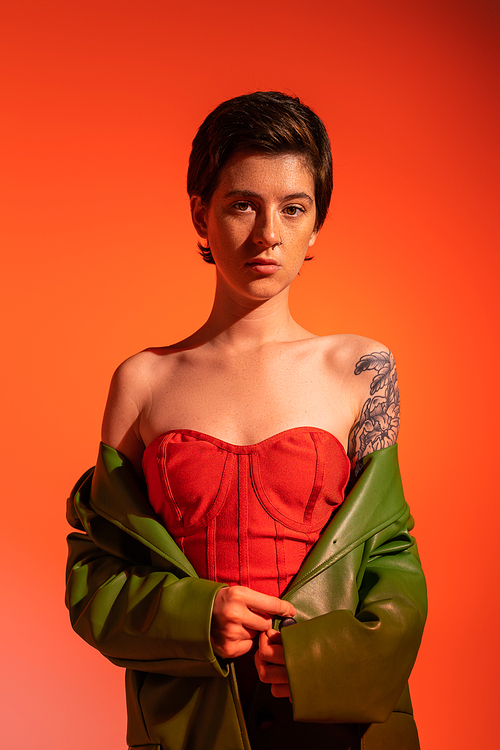young tattooed woman in red corset dress and green leather jacket looking at camera on orange background