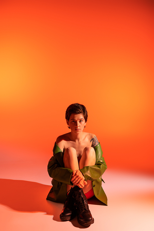 full length of brunette tattooed woman in black rough boots and green jacket sitting on orange background