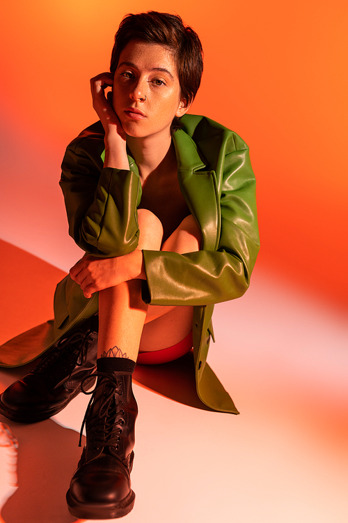 full length of dreamy woman in green leather jacket and black boots sitting and looking at camera on orange background