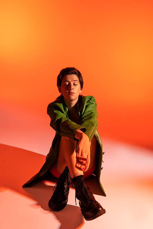 full length of pretty woman in green jacket and black boots sitting with closed eyes on orange background
