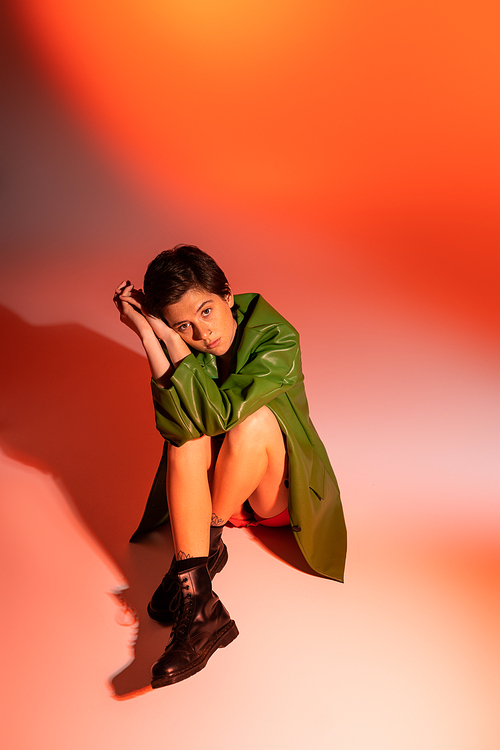trendy and pensive woman in black leather boots and green jacket sitting and looking at camera on colorful background