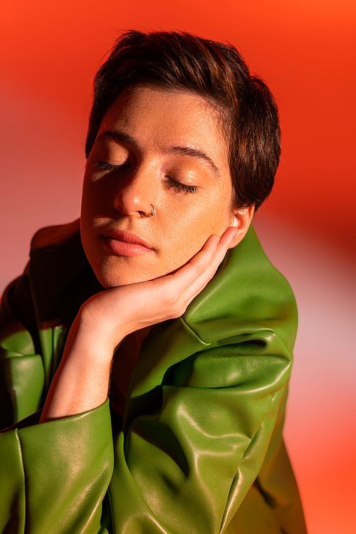 portrait of brunette woman in green leather jacket posing with closed eyes and hand near face on orange and pink background