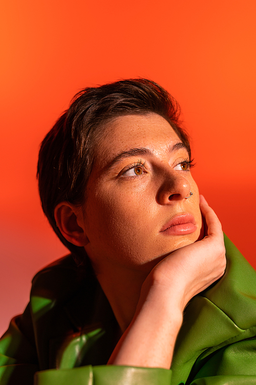 portrait of thoughtful brunette woman with freckles holding hand near face and looking away on orange background