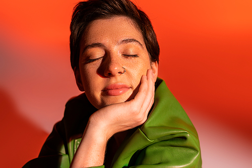 positive and dreamy woman with closed eyes holding hand near face on orange and pink background