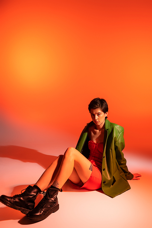 full length of fashionable brunette woman in green jacket and black leather boots sitting on orange background
